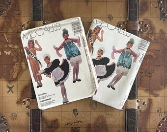May include: Two vintage McCall's sewing pattern envelopes featuring illustrations of costumes. One shows a beauty queen and a maid outfit, the other shows the same maid outfit and a blue dress. The patterns are on a brown trunk.