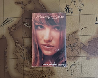 May include: A rectangular music cassette tape featuring a portrait of Britney Spears on the front cover. The text Britney Spears is printed in a white script font at the top, and the text Sneak Preview Cassette is printed in white at the bottom.