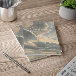 Puede incluir: Dos diarios con una portada de pintura de paisaje con un cielo nublado. La palabra "Journal" está escrita en cursiva, con el texto "My Daily" arriba y texto en alemán debajo. Un bolígrafo descansa sobre la superficie de madera.