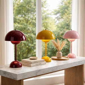 Könnte beinhalten: Drei bogenförmige Tischlampen in den Farben Burgunderrot, Gelb und Rosa. Die Lampen stehen auf einer hellen, steinähnlichen Oberfläche. Eine kleine Vase mit getrockneten Pflanzen und Bücher befinden sich ebenfalls auf der Oberfläche.