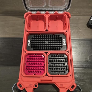 Può includere: Una custodia in plastica rossa con coperchio trasparente incernierato, dotata di scomparti interni. Contiene un vassoio nero grande con l'etichetta 9MM, un vassoio rosa piccolo con l'etichetta 9MM e un vassoio nero con l'etichetta 9MM.