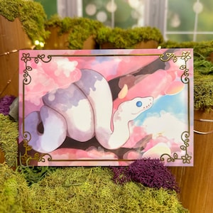 May include: A rectangular art print featuring a white and lavender snake with blue eyes, coiled around a branch. The background is a soft pink and blue sky with floral accents, framed by a gold border with floral designs.