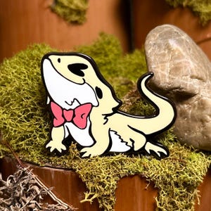 May include: A yellow and white enamel pin of a cartoon bearded dragon wearing a pink bow tie. The pin has a black outline and is set against a background of green moss and a wooden surface.