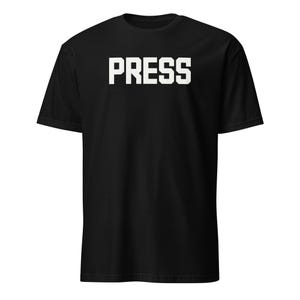 Puede incluir: Camiseta negra con la palabra "PRESS" impresa en letras blancas grandes en el pecho. La camiseta tiene mangas cortas y cuello redondo.