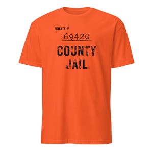 Op de afbeelding: Oranje T-shirt met de zwarte tekst "INMATE # 69420 COUNTY JAIL". Het shirt heeft korte mouwen en een ronde hals.