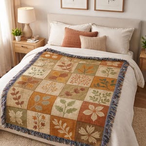May include: A woven throw blanket with a patchwork design featuring various leaf patterns in shades of brown, orange, and green. The blanket has a fringed edge and is displayed on a bed with pillows and a neutral-coloured rug.