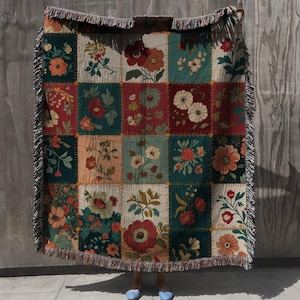 Op de afbeelding: Een patchwork plaid met bloemmotieven in rood, groen en beige tinten. De deken heeft een gefranjerde rand. De bloemmotieven zijn gerangschikt in vierkante blokken. De achtergrond is een grijze muur.