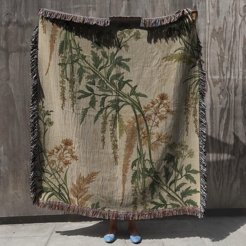 May include: A beige woven throw blanket with a botanical print featuring green and brown floral and leaf designs. The blanket has a fringed edge in shades of black, brown, and red. The blanket is held up in front of a person.