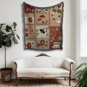 May include: A woven throw blanket with a patchwork design featuring strawberries, mushrooms, bees, and teacups. The blanket has a fringed edge and is displayed on a wall above a white sofa. The color palette includes red, beige, and green.