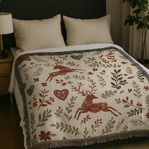 Könnte beinhalten: Beige Decke mit einem sich wiederholenden Muster aus laufenden Hirschen, Herzen und Blättern in Rot und Grau. Die Decke hat einen Fransenrand und liegt auf einem Bett mit beigen Kissen.