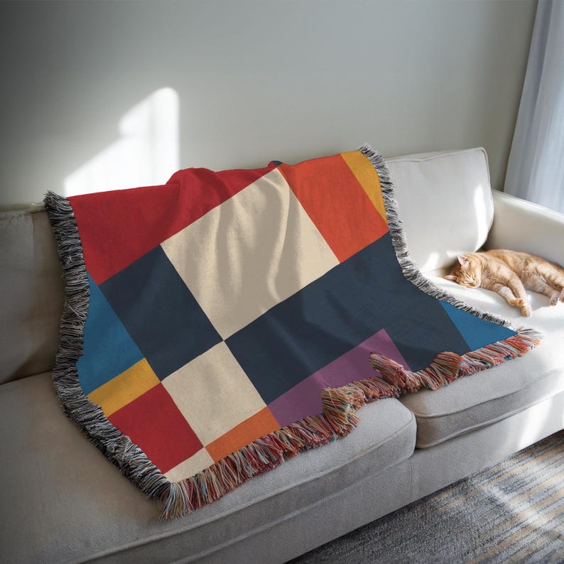 May include: A colorful throw blanket with a geometric pattern of squares in red, cream, navy, orange, yellow, blue, and purple. The blanket has a fringed edge and is draped over a white sofa. A ginger cat is sleeping on the sofa.