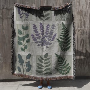 May include: A woven throw blanket with a botanical design, featuring lavender, fern, and eucalyptus illustrations in green and purple. The blanket has a fringed edge and is displayed against a wooden background.