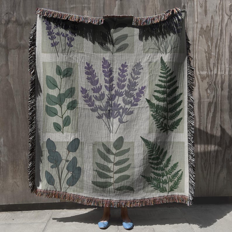 May include: A woven throw blanket with a botanical design, featuring lavender, fern, and eucalyptus illustrations in green and purple. The blanket has a fringed edge and is displayed against a wooden background.
