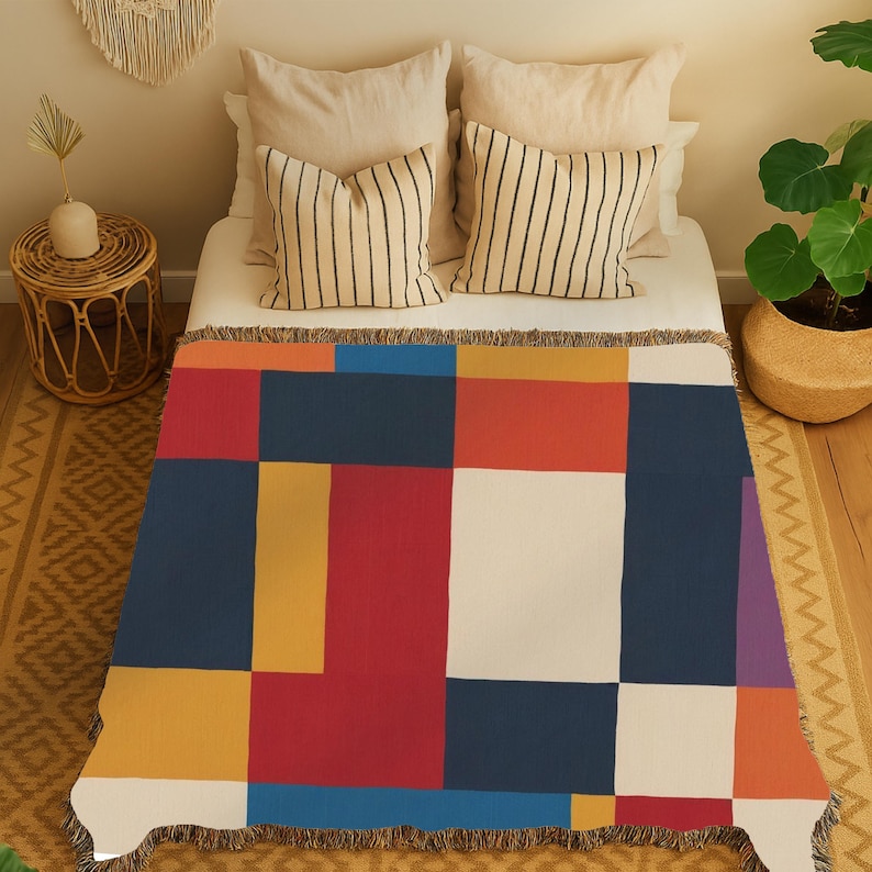 May include: A colorful throw blanket with a geometric pattern of large squares in red, blue, yellow, white, and orange. The blanket is fringed and placed on a bed with striped pillows. The overall theme is modern and abstract.