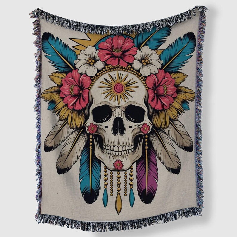May include: A beige throw blanket featuring a detailed illustration of a skull adorned with a floral headdress and feathers. The design incorporates pink, white, yellow, blue, and purple elements, with a fringed border.