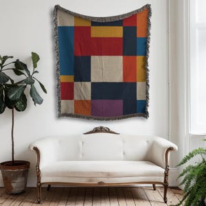 May include: A colorful woven throw blanket with a geometric pattern of rectangles in shades of red, blue, yellow, and purple. The blanket has a fringed edge and is displayed on a white wall above a white sofa.