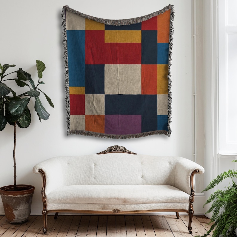 May include: A colorful woven throw blanket with a geometric pattern of rectangles in shades of red, blue, yellow, and purple. The blanket has a fringed edge and is displayed on a white wall above a white sofa.