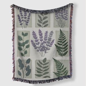 May include: A woven throw blanket with a botanical design. The design features illustrations of lavender, ferns, and other foliage in shades of purple and green, set against a light grey background. The blanket has a fringed edge.