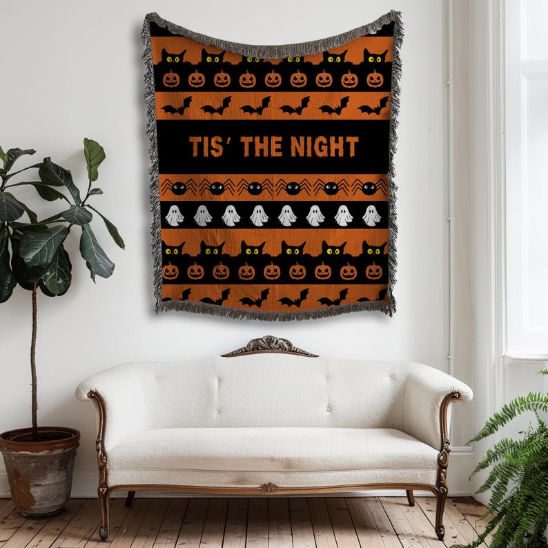 May include: Halloween-themed woven throw blanket with a black and orange pattern. The design includes black cats, jack-o'-lanterns, bats, and ghosts. The phrase "TIS' THE NIGHT" is displayed in white text on a black stripe.