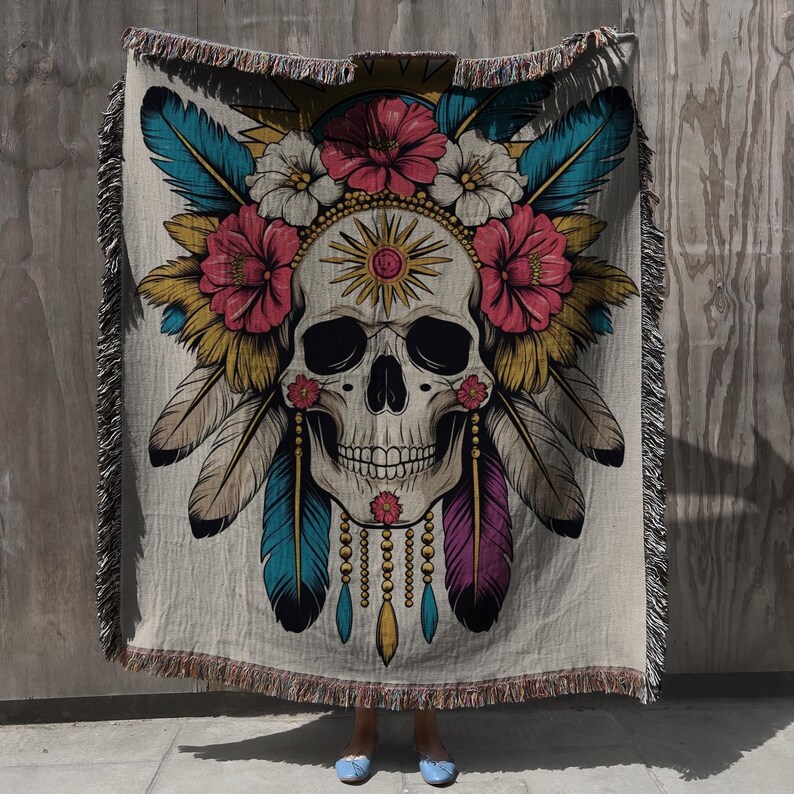 May include: A beige throw blanket featuring a detailed skull design adorned with flowers, feathers, and a sunburst. The skull is decorated with pink, white, yellow, teal, and purple accents. The blanket has a fringed edge.