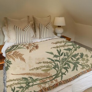 May include: A bed with a cream-colored floral throw blanket and striped pillows. The blanket features a botanical design in shades of green, brown, and beige. The pillows have vertical stripes in a dark color. A white lamp sits on a wooden nightstand.
