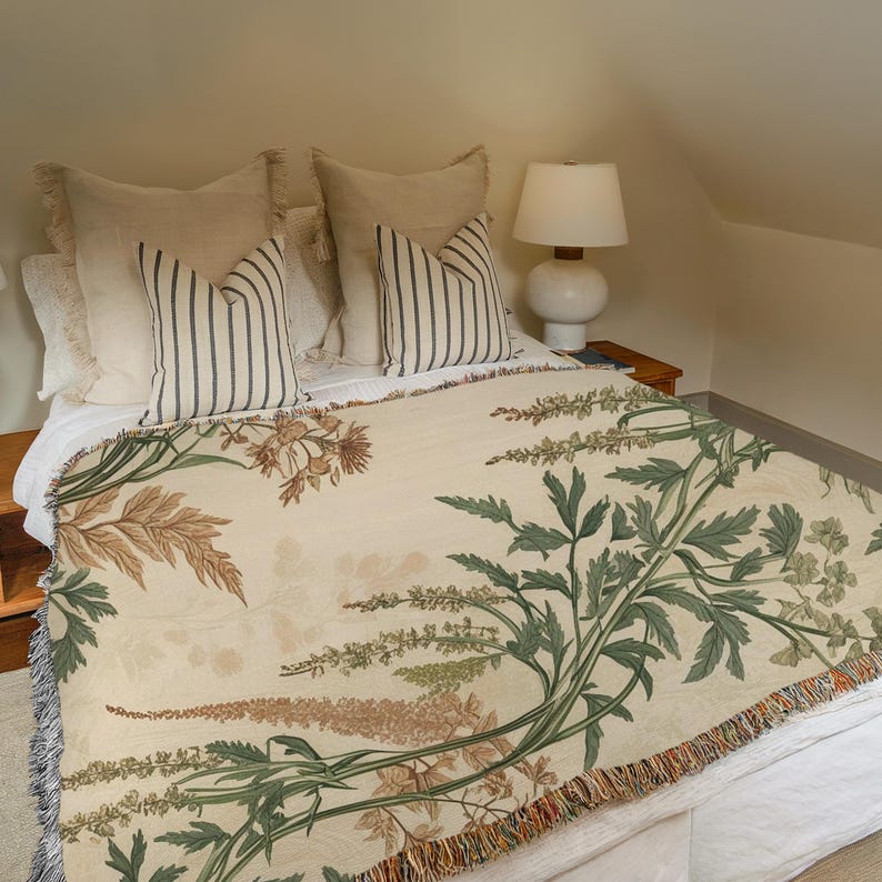 May include: A bed with a cream-colored floral throw blanket and striped pillows. The blanket features a botanical design in shades of green, brown, and beige. The pillows have vertical stripes in a dark color. A white lamp sits on a wooden nightstand.