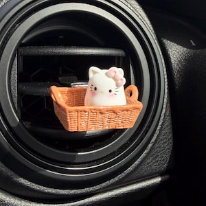 May include: A white cat figurine with a pink bow sits in a small, brown woven basket. The basket is placed in a black car air vent. The cat has black eyes and whiskers. The air vent is surrounded by a black dashboard.