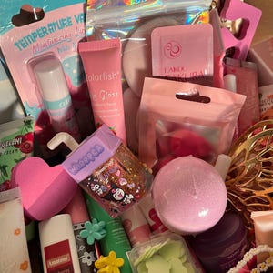 Op de afbeelding: Een doos vol diverse schoonheidsproducten, waaronder lipgloss, lippenbalsem en een badbruisbal. De items zijn voornamelijk roze en pastelkleurig, met teksten als "Colorfish" en "Amatoni". Een Hello Kitty-tas is ook zichtbaar.