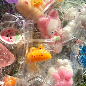 May include: A collection of colorful, small, plastic toys in clear plastic bags. The toys include a pink bunny, a yellow frog, a white dog, a purple blob, and a pink paw print. Some toys have glitter accents.