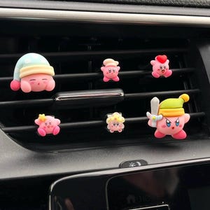 Puede incluir: Colección de adornos de ventilación para coche de color rosa, estilo dibujos animados. Los adornos presentan varios diseños, incluyendo un personaje con gorro de dormir, gorro de chef, corazón, estrella, flor y espada. Los adornos están fijados a la rejilla de ventilación de un coche.