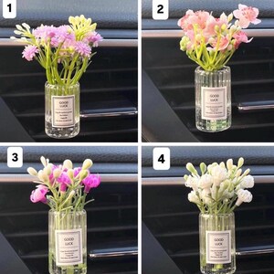 May include: Four car air vent diffusers, each with a different arrangement of artificial flowers in a clear, ribbed glass bottle. Each bottle has a label that reads "GOOD LUCK". The flowers are pink, white, and yellow.