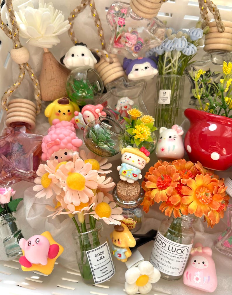 Op de afbeelding: Een verzameling decoratieve luchtverfrissers en miniatuur bloemstukken. De collectie bevat stripfiguren, kleine glazen flesjes en kunstbloemen in verschillende kleuren, waaronder roze, geel en oranje. Sommige flesjes hebben de tekst "GOOD LUCK".