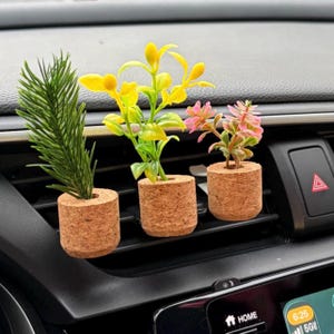 Puede incluir: Tres pequeñas plantas artificiales en macetas de corcho. Las plantas presentan hojas verdes, amarillas y rosas. Las macetas son cilíndricas y están colocadas en una rejilla de ventilación del coche.