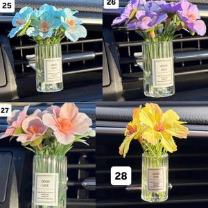 May include: Four small glass vases, each containing a different color of artificial flowers. The flowers are blue, purple, pink, and yellow. Each vase has a label that reads "GOOD LUCK". The vases are designed to be placed in a car's air vent.
