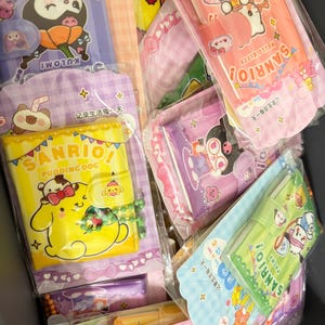 Op de afbeelding: Een verzameling Sanrio-thema briefpapier, waaronder notitieboekjes en etuis, in verschillende kleuren zoals roze, geel en paars. De items bevatten stripfiguren en tekst, allemaal verpakt in doorzichtig plastic.