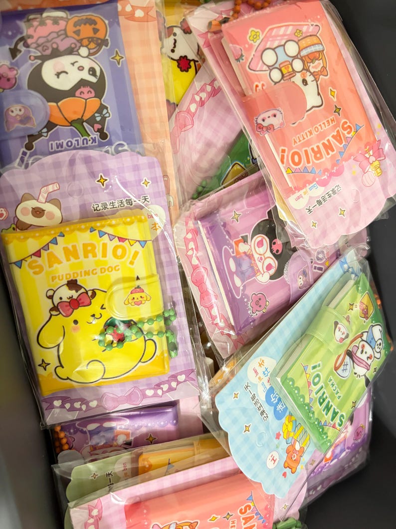 Op de afbeelding: Een verzameling Sanrio-thema briefpapier, waaronder notitieboekjes en etuis, in verschillende kleuren zoals roze, geel en paars. De items bevatten stripfiguren en tekst, allemaal verpakt in doorzichtig plastic.