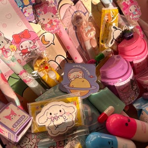 Op de afbeelding: Een verzameling kleurrijke briefpapier en accessoires, waaronder pennen, gummen, stickers en een paarse beker. De items bevatten stripfiguren en ontwerpen, met roze, blauwe en gele accenten. Het woord "universe" is zichtbaar op een sticker.