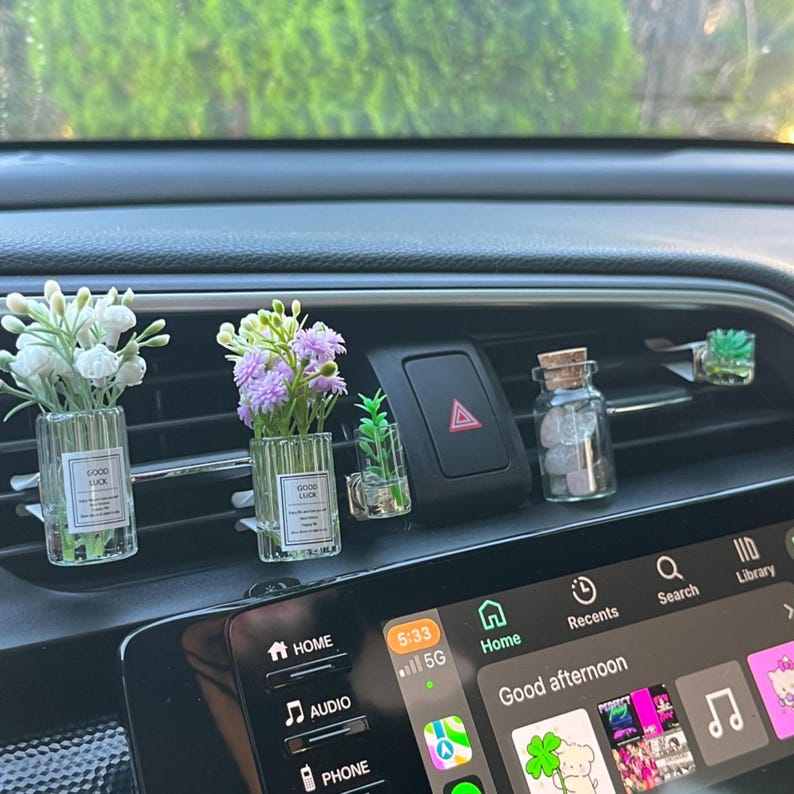 May include: A car air vent featuring several decorative items. There are small glass vases with white and purple flowers, a tiny plant, and a glass jar with a cork lid filled with stones. The dashboard also displays a digital screen.