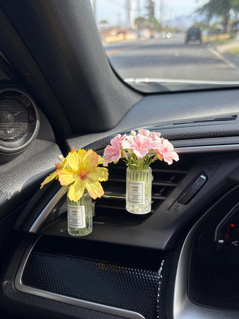 May include: Two small glass car air fresheners with floral arrangements. One has yellow flowers, the other pink. Each has a label that reads "GOOD LUCK". The fresheners are clipped to the car's air vents.