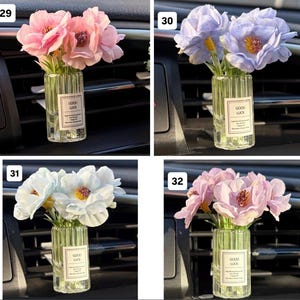 May include: A small glass vase with a label that reads "GOOD LUCK" contains a bouquet of pink flowers. The vase is placed in a car's air vent. The flowers have a realistic appearance.