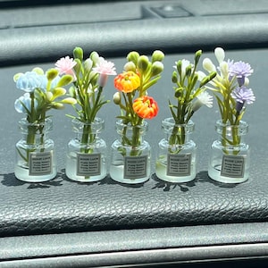 Könnte beinhalten: Fünf kleine Glasvasen, jede mit einer anderen Anordnung von bunten Kunstblumen. Die Vasen haben ein Etikett mit der Aufschrift "GOOD LUCK Pretty Flowers". Die Blumen umfassen blaue, rosa, orange, weiße und lila Blüten.