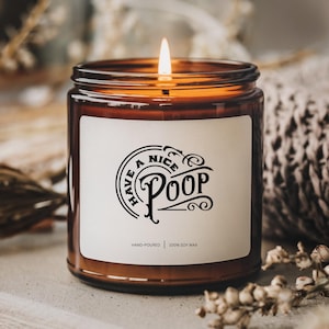 May include: A lit brown glass candle jar with a white label. The label features the phrase "Have a Nice Poop" in a black, decorative font. The candle is hand-poured and made with 100% soy wax.