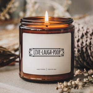 May include: A lit brown glass candle jar with a white label featuring the text "LIVE.LAUGH.POOP" in black. The candle is hand-poured with 100% soy wax. The flame is lit, and the background is blurred.