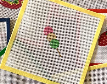May include: Needlepoint canvas with a yellow border featuring a colorful design of a dango skewer. The dango has a pink, green, and white color scheme. Other needlepoint canvases are visible in the background.