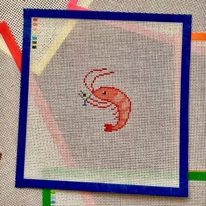 May include: A square needlepoint canvas featuring a white grid background and a blue fabric border. In the center, a small orange shrimp is depicted holding a tiny martini glass. A color key with small squares is printed in the top left corner.