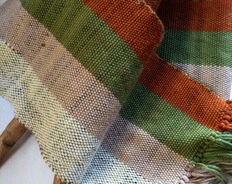 May include: A woven scarf with stripes of beige, green, and orange. The scarf has fringe on the ends and a label that reads "Handwoven by Diana Miller Backstrap Loom".