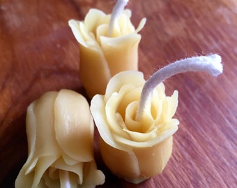 May include: Three small, yellow, wax candles shaped like roses with white wicks. The candles are sitting on a wooden surface.