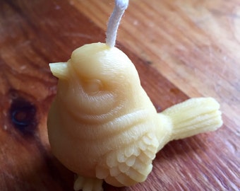 May include: A small, yellow beeswax candle shaped like a bird with a white wick. The candle has detailed features, including a beak, eyes, and feathers.