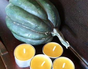 May include: Four round, yellow tea light candles in metal holders, sitting on a dark brown wooden surface next to a green acorn squash.