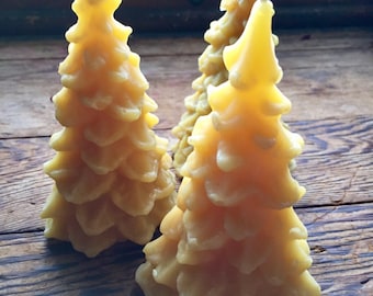 May include: Three yellow beeswax candles shaped like Christmas trees. The candles are on a wooden surface.